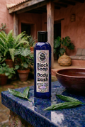 Cobalt-blue Black Soap Body Wash bottle on wet sapphire granite beside a burnished clay bowl in a reddish earthen courtyard.